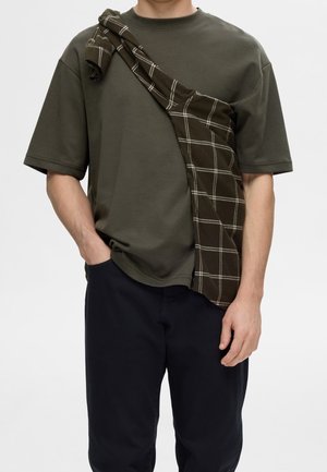 Personne portant une chemise à manches courtes vert foncé avec une chemise à carreaux marron drapée sur une épaule et un pantalon noir, main dans la poche.