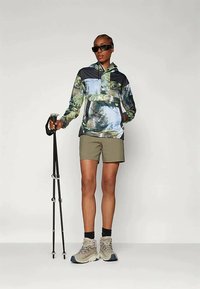 Vandringsoutfit med en flerfärgad, mönstrad tröja med huva, beige shorts och lätta vandringsskor; håller en vandringsstav.