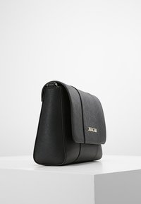 Borsa a tracolla nera in pelle texturizzata con chiusura a patta e discreto logo metallico sul davanti, esposta su una superficie bianca.