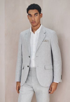 Mand iført lysegråt jakkesæt med hvid skjorte og beige lommetørklæde, stående mod en ensfarvet beige baggrund.