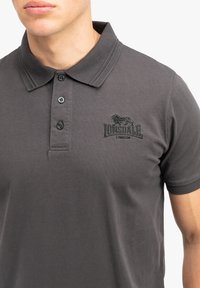 Šedé bavlněné polo tričko se strukturovaným límcem, zapínáním na tři knoflíky a vyšitým logem na levé hrudi. Krátké rukávy.