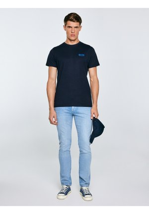 Noor mees tumesinises "Big Star" T-särgis, helesinistes teksades, tumelillades tossudes, hoides tumelillat nokamütsi ühevärvilise halli tausta ees.