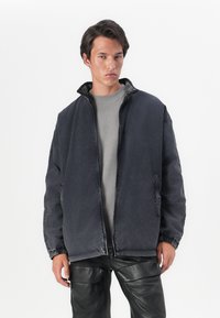 Jaqueta de ganga escura com colarinho alto, apresentando punhos elásticos e bolsos laterais, combinada com um suéter cinza de gola redonda e calças de couro pretas.