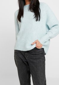 Maglione azzurro chiaro a maglia con una texture a coste, maniche larghe e un orlo leggermente curvo, abbinato a jeans grigio scuro.