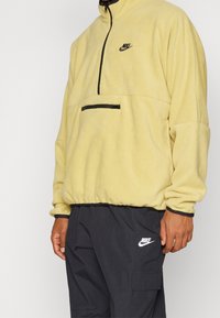 Gul fleece-pullover med halv dragkedja fram, svart dragkedja och bröstficka. Har en liten svart Nike-logotyp på bröstet och byxorna.