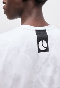 Κοντινή λήψη του λαιμού και του άνω μέρους της πλάτης ενός ατόμου που φοράει λευκό πουκάμισο με διακριτικά κυκλικά σχέδια και μια μαύρη ετικέτα με ένα λευκό εικονίδιο τένις.