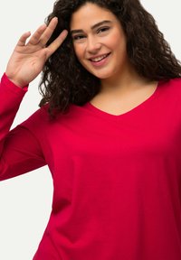 Langarm V-Ausschnitt-Oberteil aus leuchtend pinkem Stoff. Glatte Textur, figurschmeichelnde Passform mit lockerer Silhouette. Das Model winkt und zeigt die Vielseitigkeit des Oberteils.