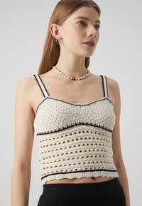 Top in cotone all'uncinetto di colore bianco sporco con rifiniture nere. Caratterizzato da un corpo aderente e un motivo di cucitura aperta. Decorato con bordi a onde.