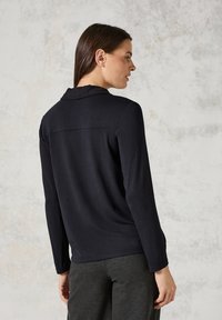 Maglietta nera a maniche lunghe con colletto, caratterizzata da una texture liscia e una vestibilità leggermente affusolata, abbinata a pantaloni grigi a gamba affusolata.