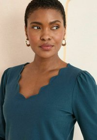 Blouse bleu-vert avec un col festonné et des manches bouffantes. Tissu lisse avec un éclat subtil. De grandes boucles d'oreilles en or rondes mettent en valeur le look.