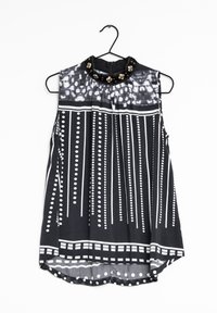 Blusa sin mangas en blanco y negro con un cuello alto, que presenta un diseño plisado, patrones circulares y tachuelas decorativas en el escote.