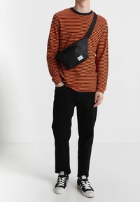 Černá crossbody taška s přední kapsou na zip, nošená přes oranžovo-černé pruhované tričko s dlouhým rukávem, v kombinaci s černými kalhotami a teniskami.