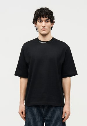 Jeune homme aux cheveux bouclés foncés portant un T-shirt noir oversize avec le logo "Les Benjamins" au col et un pantalon à fines rayures, debout de manière neutre.