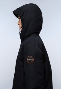 Černá kapucová bunda vyrobená z texturované látky, s kulatou nášivkou na levém rukávu a nastavitelnou šňůrkou na kapuci.