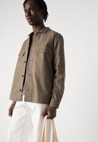Light brown button-up jacket with two chest pockets, worn over a gray top. Model holds a beige tote bag, paired with white pants.