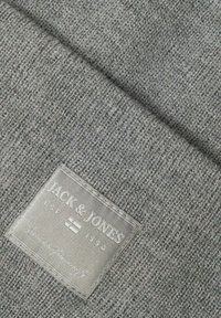 Grå strikket stof med en tekstureret overflade, featuring et syet sølvmærke, der inkluderer "JACK & JONES" og "EST. 1990."