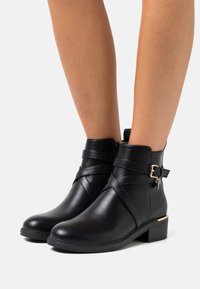 Bottines noires avec une finition lisse, dotées de sangles croisées, d'une boucle sur le côté et d'un petit accent doré sur le talon. Panneau latéral texturé.