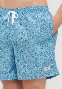 Sinine mustriga ujumispüksid elastse vöökohtaga, valge nööriga, küljetaskudega ja väikese logoplaastriga alumisel jalal.