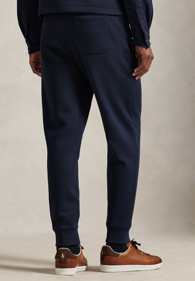 Στενά μπλε παντελόνια φόρμας, κατασκευασμένα από απαλό ύφασμα, με πίσω τσέπη και ριμπ μανσέτες. Φαίνονται καφέ αθλητικά παπούτσια με λευκή σόλα.