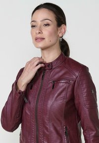 Veste en cuir bourgogne avec un col montant, fermeture éclair à l'avant et détails de panneaux, dotée de plusieurs poches zippées et d'une texture lisse.