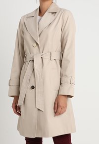 Beige trenchcoat med dubbla knappar, bred krage och bälte. Har stora knappar och texturerade ärmdekorationer, knä-lång.