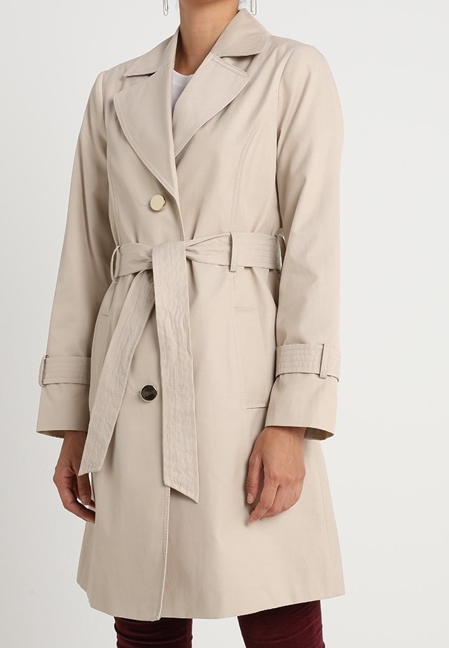 Beige trenchcoat med dubbla knappar, bred krage och bälte. Har stora knappar och texturerade ärmdekorationer, knä-lång.