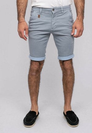 Homme portant un short bleu clair retroussé, chemise blanche, chaussures noires sans lacets, debout les bras détendus le long du corps sur fond gris.