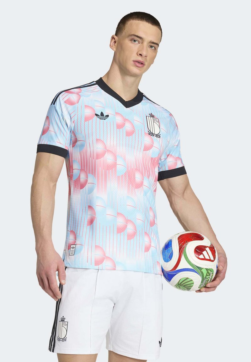 Mand iført pastelblå og lyserøde mønstrede fodboldtrøje med sort kant, hvide shorts, der holder en rød, blå, grøn og hvid fodbold.