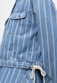 Μπλε τζιν μπουφάν με λευκές κάθετες ρίγες, πατιλέτα τσέπης στο στήθος και λευκό κορδόνι δεμένο στη μέση.