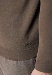 Maglietta a maniche lunghe in maglia marrone con orlo e polsini a coste. Presenta un piccolo etichetta con il marchio sul lato. Texture liscia, design aderente.