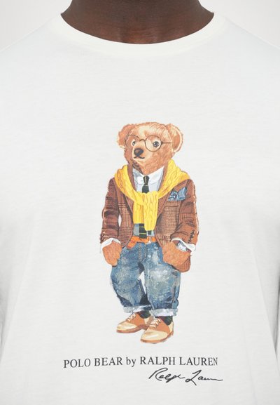 T-shirt gráfica com um urso vestido com um blazer castanho, suéter amarelo, camisa branca, óculos, jeans azuis e sapatos castanhos. Fundo branco.