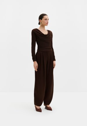 Femme debout portant un haut drapé à manches longues marron foncé, un pantalon à larges jambes, des talons pointus marron et des boucles d'oreilles dorées sur un fond uni.