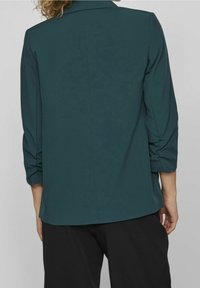 Tunique turquoise à manches longues, avec des détails froncés aux poignets et une coupe droite. Tissu doux avec une texture lisse.