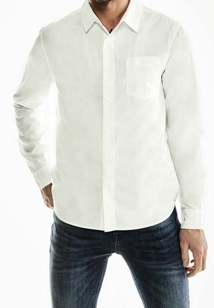 Uomo con camicia bianca a maniche lunghe con taschino e jeans blu scuro, in piedi con le mani vicino ai fianchi.