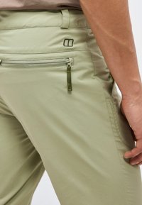 Gros plan sur un pantalon vert clair avec une poche arrière zippée et un logo noir près de la taille, porté par une personne avec le bras droit détendu le long du corps.