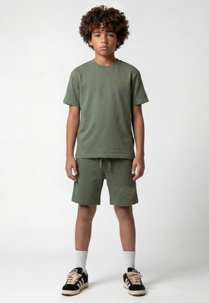 Jongen met krullend haar staat naar voren, draagt een groene t-shirt en shorts-set, witte sokken en zwarte sneakers op een effen achtergrond.