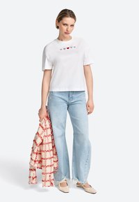 White cotton t-shirt with short sleeves, featuring five small embroidered hearts in red and blue. Paired with light blue wide-leg jeans and a red-checkered shawl.