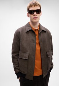 Jauns vīrietis valkā brūnu herringbone žaketi virs apdeguma oranžas polo krekla un tumšiem saulesbrillēm, stāvot ar rokām kabatās.
