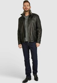 Chaqueta de piel negra con cuello de peluche, bolsillos con cremallera, llevada sobre una camiseta henley gris y jeans oscuros, combinada con zapatos negros.