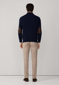 Mann trägt einen marineblauen Pullover mit braunen Ellbogenpatches, beigen Hosen und braunen Schuhen und steht vor einem einfarbigen hellgrauen Hintergrund.