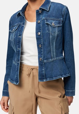 Mujer con una chaqueta vaquera azul ajustada sobre una camiseta blanca, combinada con pantalones cargo beige con cordón, de pie frente a un fondo liso.