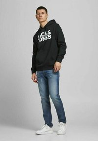 Sudadera negra con gran logo blanco "JACK & JONES", bolsillo frontal, combinada con jeans azules desgastados y zapatillas blancas, en pose de pie.