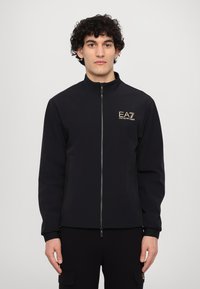 Svart zip-up jacka med hög krage, med en gyllene logga på bröstet och sidofickor. Slättyg med en figursydd design.