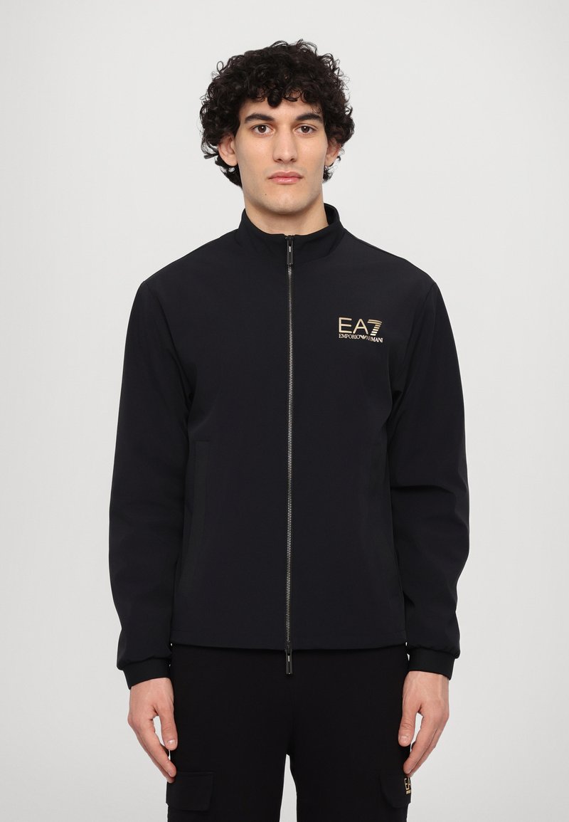 Svart zip-up jacka med hög krage, med en gyllene logga på bröstet och sidofickor. Slättyg med en figursydd design.