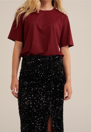 Vrouw draagt een bordeaux kortemouwen shirt en een zwarte pailletten rok met een vooraan een split, staand tegen een effen lichte achtergrond.