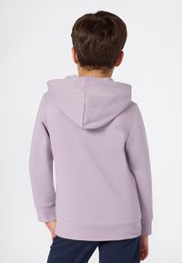 Enfant portant un sweat à capuche violet clair et un pantalon bleu marine, vu de dos sur un fond blanc uni.