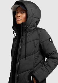 Chaqueta de plumas negra con capucha, que presenta un cierre frontal con cremallera, cordones ajustables y un patrón acolchado. Incluye un acento de marca en la manga.