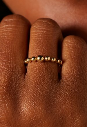 Gouden ring met een rij van gladde kralen in verschillende maten, tegen een textuur van bruin huid, die het reflecterende oppervlak benadrukt.