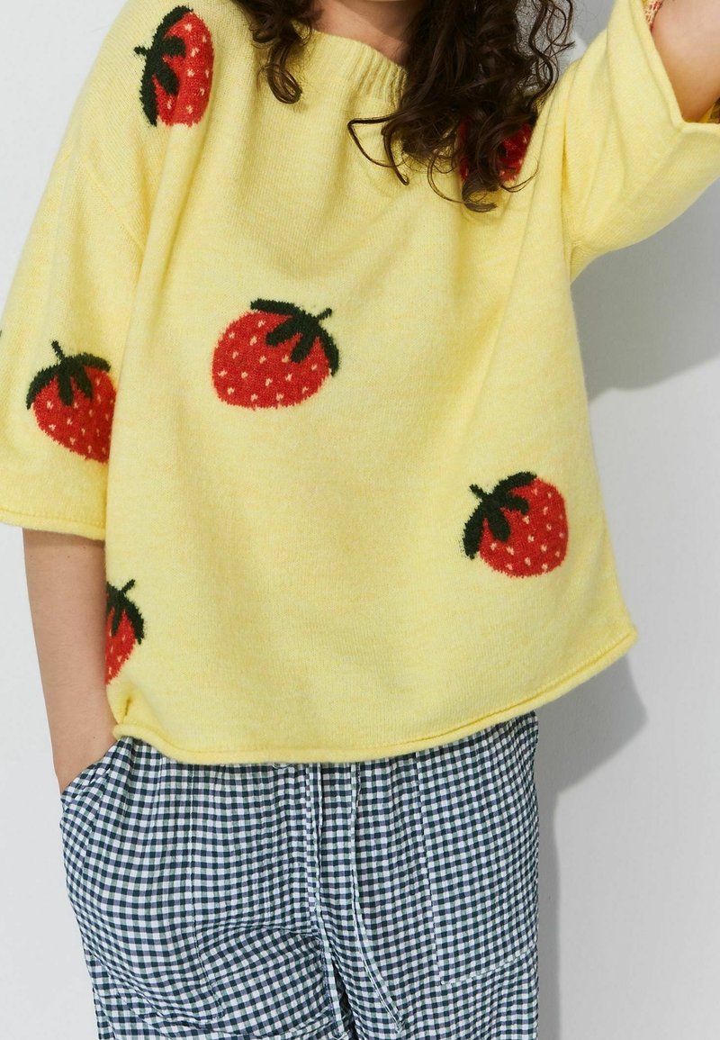 Personne portant un haut en tricot jaune avec des fraises rouges et un pantalon à carreaux bleu et blanc, main dans la poche, bras levé.