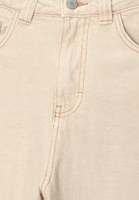 Gros plan sur un pantalon en denim beige montrant le bouton avant, les passants de ceinture et les coutures détaillées autour de la poche et de la zone de la fermeture éclair.